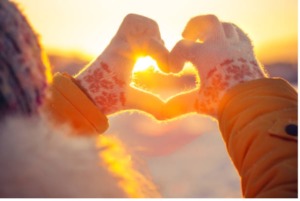 Picture of person wearing snowflake gloves; using index fingers and thumbs to make a heart framing the sunset in the background