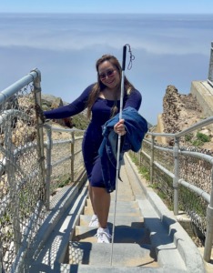 Photo of Sweet Ann Carol Nanca posing with her white cane on a stairway to the beach