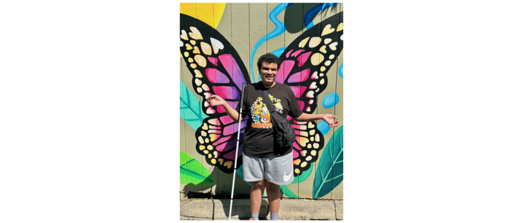 Banner photo of Charlie Turner smiling and posing in front of a butterfly mural appearing like the wings are coming out of his back.